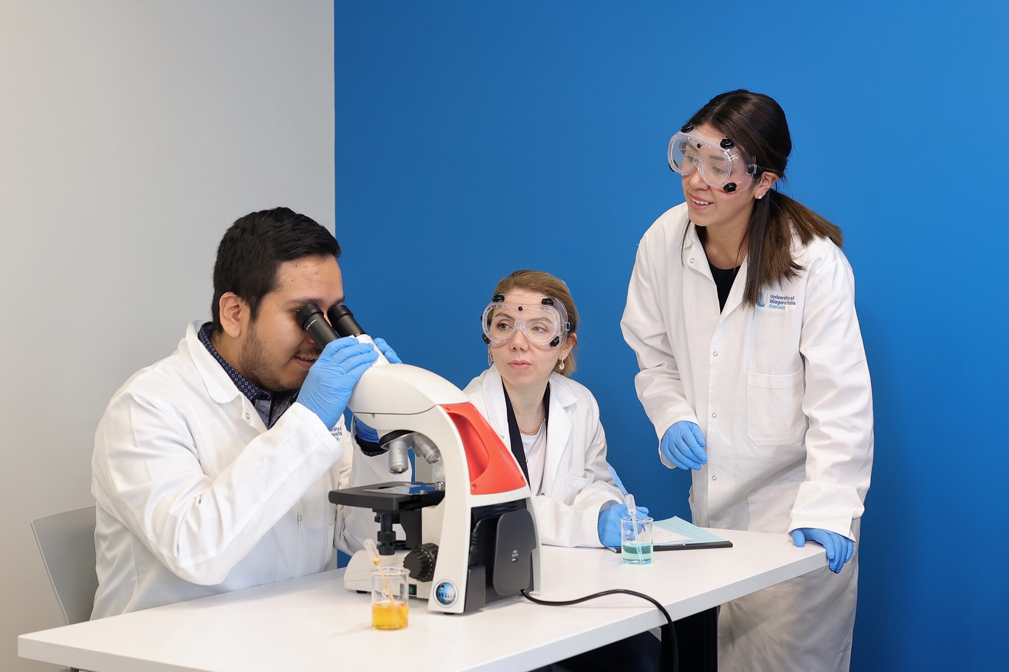 Three individuals in lab coats examining a microscope, engaged in scientific research and analysis in a laboratory setting