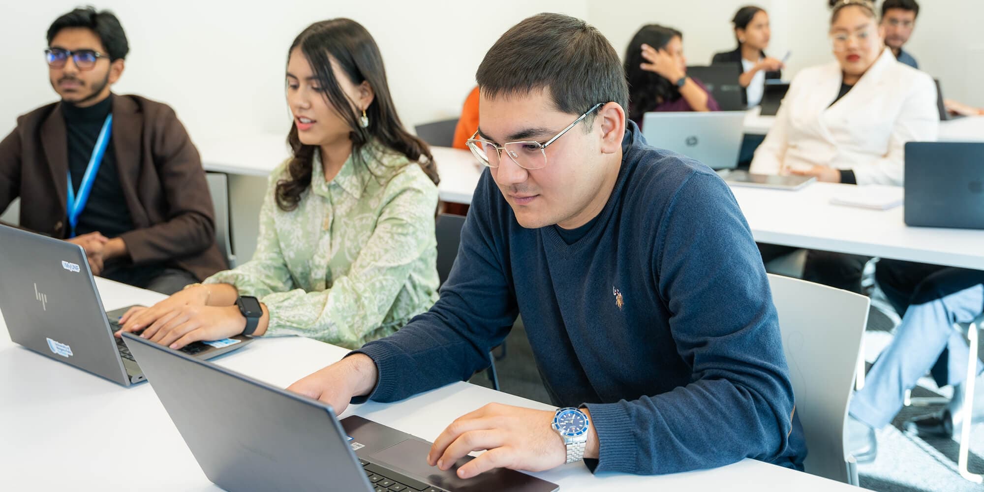 A diverse group of individuals seated at tables, each engaged with their laptops in a collaborative environment