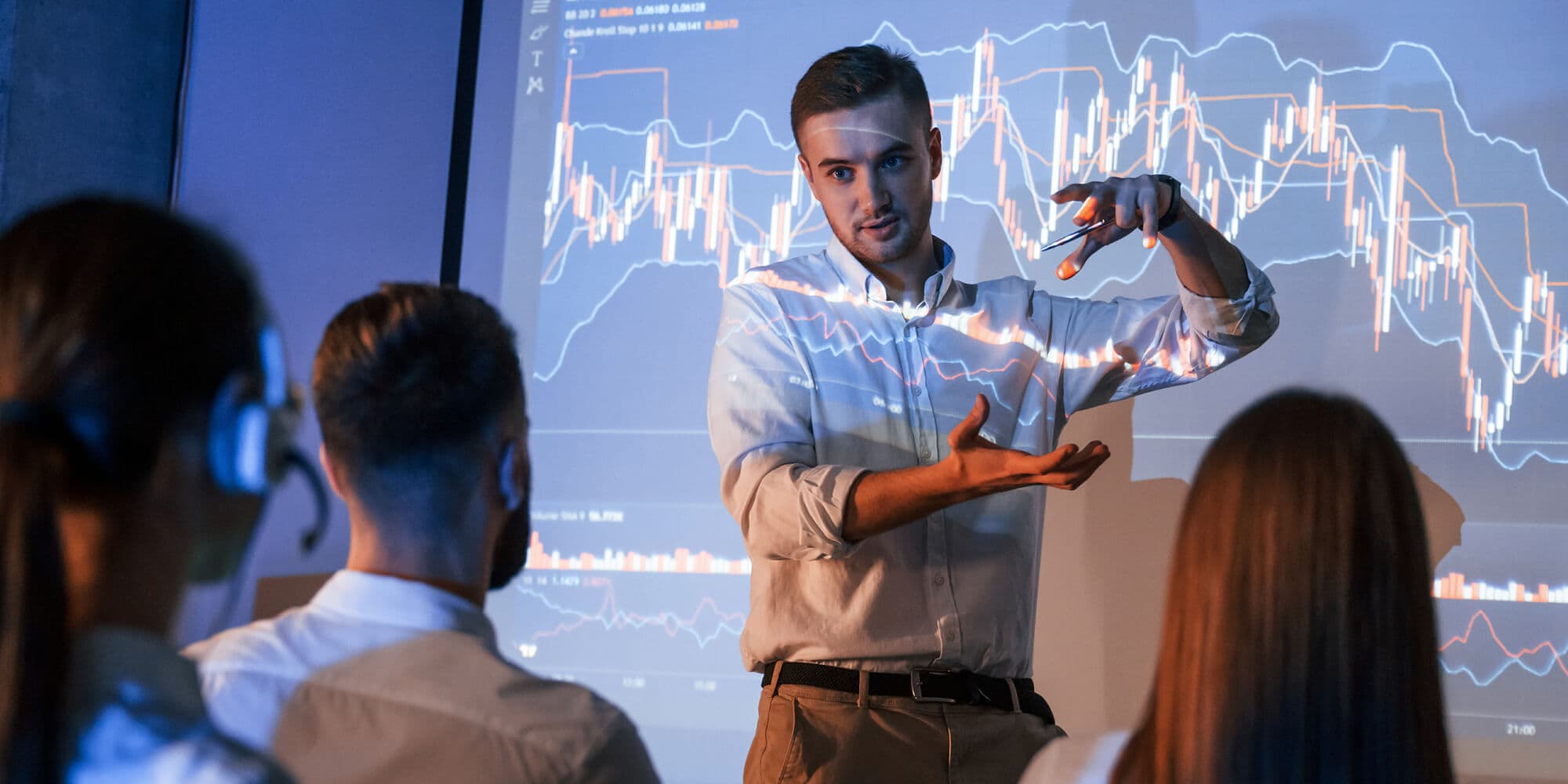 A man stands before a screen displaying a stock market chart, discussing data