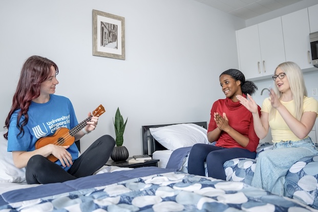 Three people are sitting on beds in a dorm room, one playing a ukulele while the other two clap along.