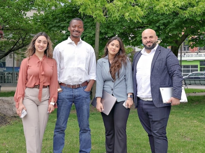 Four individuals stand together in a park, smiling and posing for a group photo amidst greenery and trees.