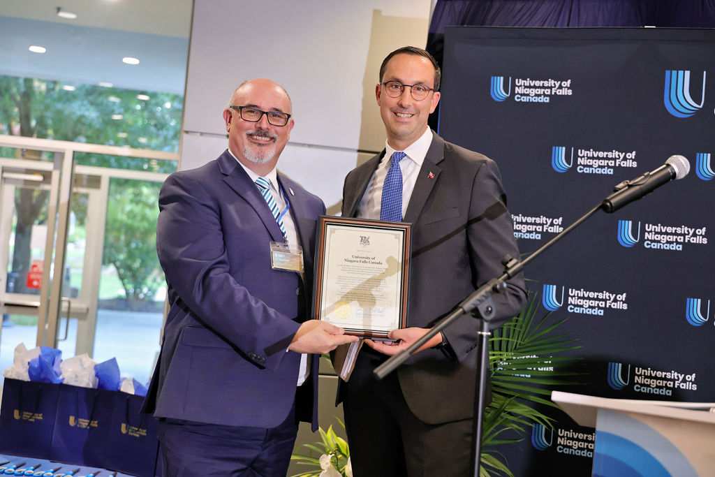 Two men in formal suits and ties proudly holding an award