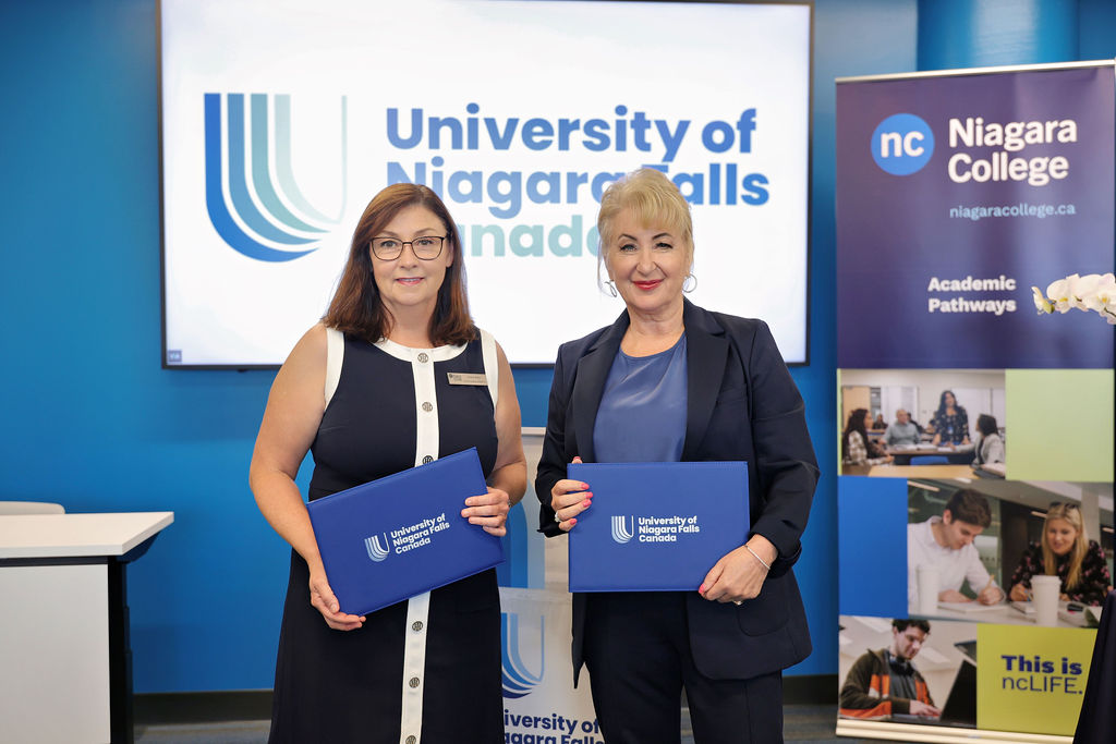 Two women display blue papers in front of a blue sign, emphasizing their shared message and collaboration