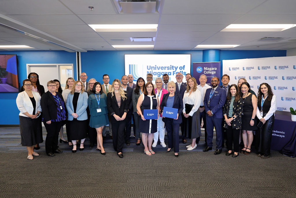 A diverse group of individuals stands together in front of a large blue banner, smiling and engaged in conversation.