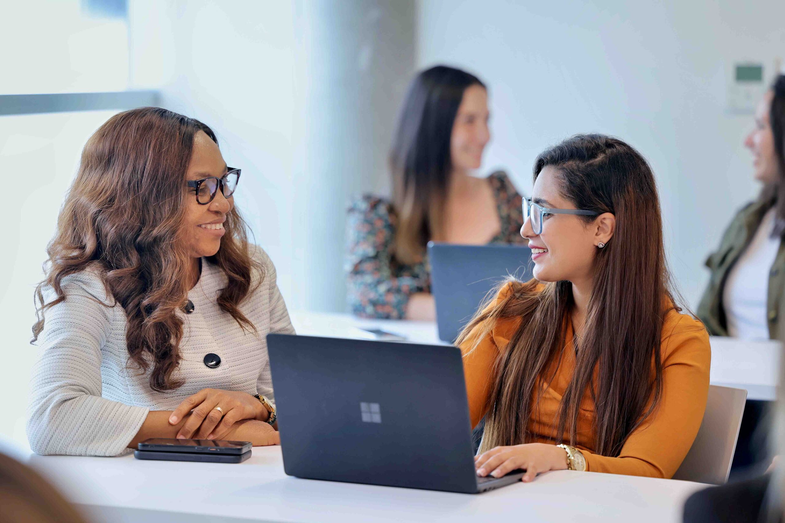 Two women engaged in work, sitting at a table with laptops, focused on their tasks in a collaborative environment.